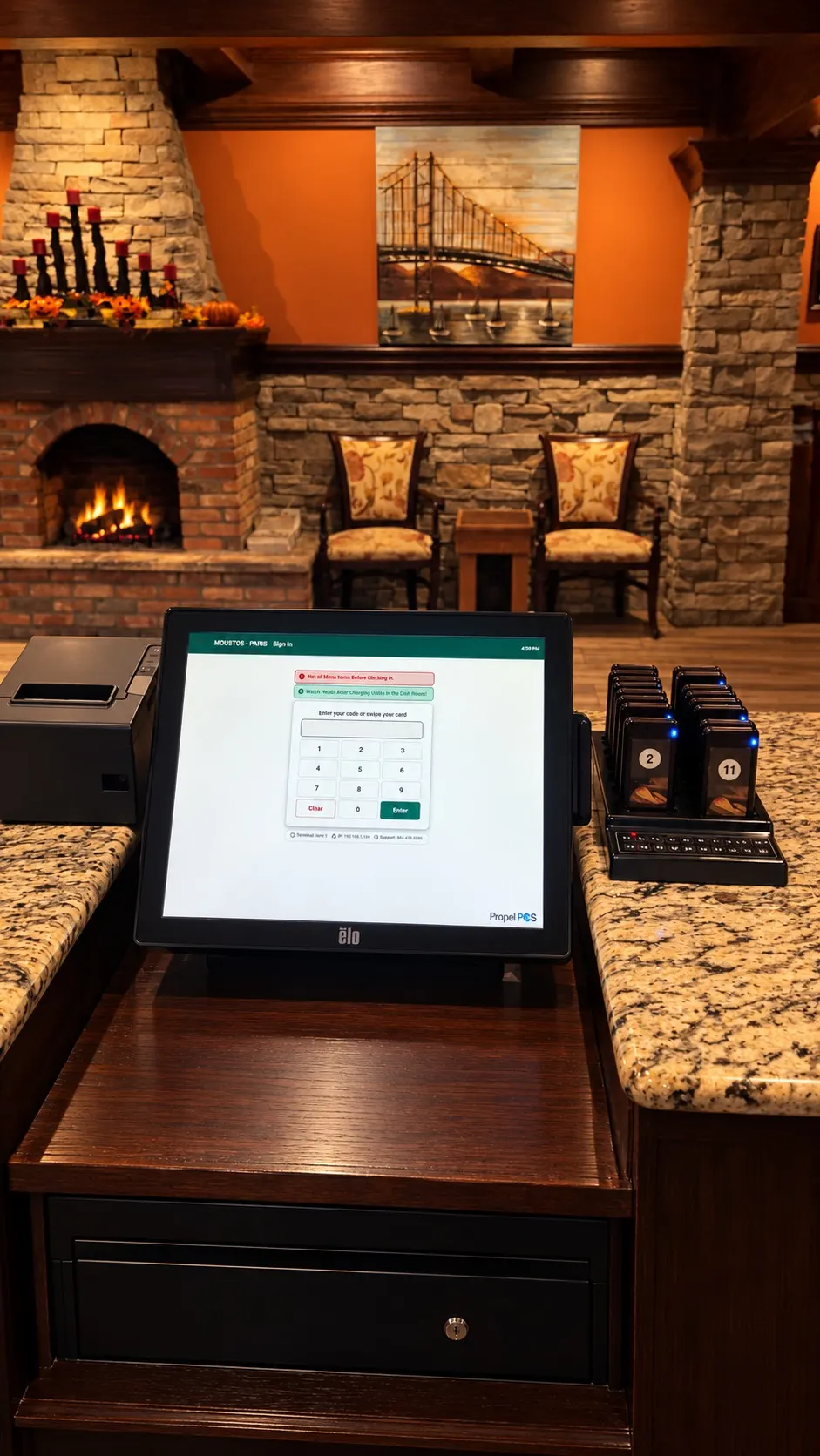 A Propel POS touchscreen station on the front counter at Moustos Pasta & Grill in Paris, Tennessee, with a receipt printer, guest pagers and a stone fireplace in the dining room behind it