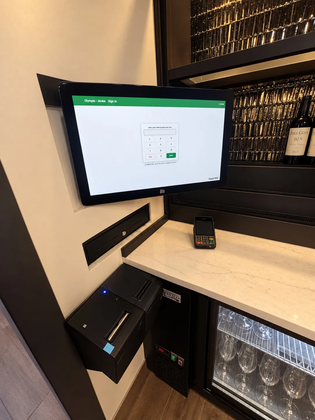 A Propel POS touchscreen station on a swing arm at Olympic Steak & Pizza in Atoka, Tennessee, showing the sign-in keypad, with a cash drawer built into the wall, a card reader on the marble bar top, receipt printers below and a glass-door cooler of wine glasses